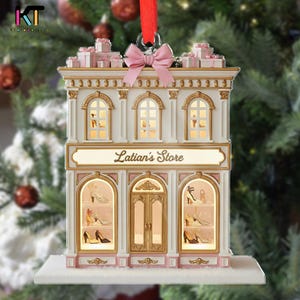 May include: A Christmas ornament shaped like a store, featuring the text "Latian's Store". The building is white with gold trim and pink accents, with lit windows displaying shoes. A pink bow and small gift boxes adorn the top.