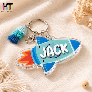 May include: A rocket-shaped acrylic keychain featuring the name JACK in white raised lettering. The rocket is light blue with dark blue accents and a red flame at the base. It includes a silver metal keyring, a lobster clasp, and a three-tiered tassel in shades of light blue, teal, and dark blue.