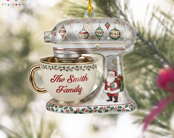 May include: A Christmas ornament shaped like a silver stand mixer with a gold-trimmed cup. The cup reads "The Smith Family" in red script. The mixer is decorated with ornaments and a Santa Claus figure.