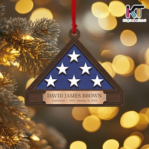 May include: A wooden ornament shaped like a folded American flag with a blue field of stars and a brown wooden frame. The ornament has a brass plaque with the text "DAVID JAMES BROWN September 1, 1995 - January 18, 2024".