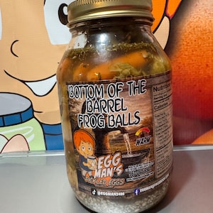 May include: A glass jar filled with pickled vegetables and eggs. The jar has a gold lid and a label that reads "BOTTOM OF THE BARREL FROG BALLS" with the text "EGG MAN'S PICKLED EGGS". The jar is on a light surface.