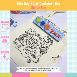 Peut inclure: Un sac fourre-tout crème avec un motif "Dino Surf" en noir et blanc. Une boîte de peinture avec des pots de peinture verts, bleus et oranges est posée dessus. Le texte "Tote Bag Paint Container Box Digital File" est visible.