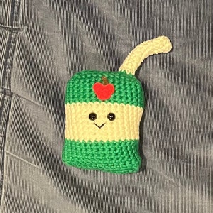 May include: A handmade crochet apple-shaped item with a smiling face, black button eyes, and a red heart-shaped apple. The item is green and cream-colored with a curved stem. The item is likely a decorative piece or a toy.