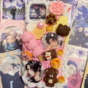 Puede incluir: Una funda de teléfono decorativa con base rosa, adornada con varios abalorios, incluyendo una huella de pata, una barra de chocolate, un oso y Hello Kitty. La funda también presenta una imagen enmarcada y el texto "umibunn".