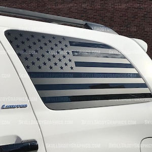 May include: A white SUV with a rear window decal featuring the American flag. The flag design includes blue stars and stripes. The decal is applied to the window of the vehicle. The text "SKULLDADDYGRAPHICS.COM" is visible.