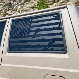 May include: A tan vehicle with a rear window featuring a distressed American flag design. The flag's stars and stripes are visible, with a weathered appearance. The vehicle has a black antenna and a dark-tinted window.