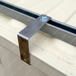 May include: Close-up of a metal bracket attached to a metal track with a screw. The bracket is rusty and has several holes. The track is silver and has a black rubber strip running along the top.
