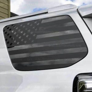 May include: A black and gray American flag graphic applied to a vehicle window. The flag design includes stars and stripes, with the text "SKULLOADDYGRAPHICS.COM" repeated across the image.