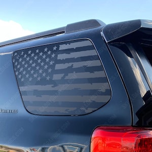May include: A black vehicle window decal featuring a distressed American flag design. The flag is rendered in shades of gray, with visible stars and stripes. The decal is applied to the rear window of a black SUV.
