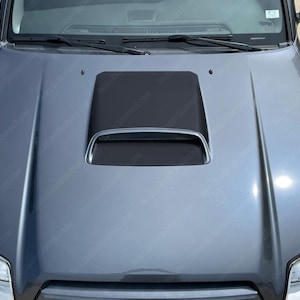 May include: A close-up shot of a gray car hood with a black hood scoop. The hood scoop has a silver trim and a mesh grille. The car's hood is clean and reflects the surrounding environment.
