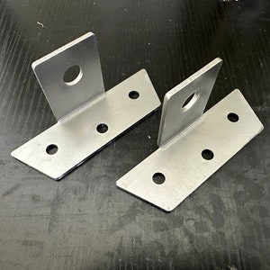 May include: Two silver metal brackets with four holes each. The brackets are angled and have a large hole in the top corner.