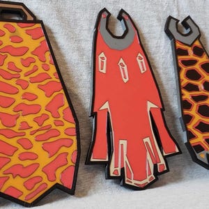 May include: Three decorative, stylized capes in shades of red, orange, and black. The capes have unique patterns and shapes, with a handle-like design at the top. The designs include a spotted pattern, geometric shapes, and a flame-like pattern.