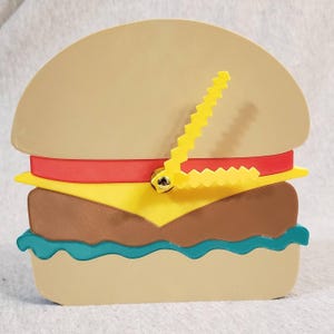 Könnte beinhalten: Eine neuartige Wanduhr in Form eines Hamburgers. Die Uhr hat ein hellbraunes Brötchen, ein rotes Band, gelben Käse, einen braunen Burger und türkisfarbenen Salat. Die Zeiger sind gelb und gezackt. Ein lustiges Deko-Objekt.