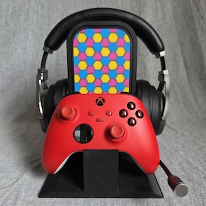 May include: A red Xbox controller rests on a black stand, with a headset and a stand featuring a colorful geometric pattern. The headset is black with silver accents. The stand is designed to hold gaming accessories.