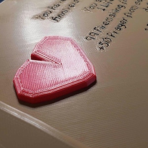 Op de afbeelding: Een rood, hartvormig object met een ontbrekend deel, rustend op een beige oppervlak. Het object heeft een getextureerd uiterlijk. Tekst is zichtbaar op de achtergrond, waaronder de woorden "Congratulations" en "Anniversary Manager."