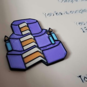 Op de afbeelding: Een paarse, drielaagse taartspeld met oranje en witte lagen en twee blauwe kaarsen. De speld heeft een zwarte omtrek en staat tegen een lichte achtergrond met tekst.