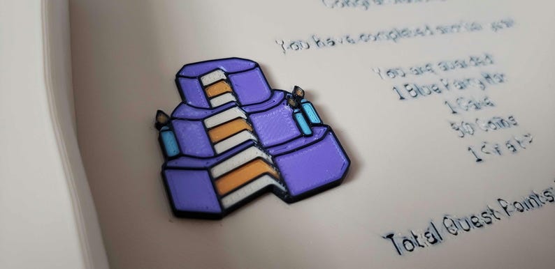 Op de afbeelding: Een paarse, drielaagse taartspeld met oranje en witte lagen en twee blauwe kaarsen. De speld heeft een zwarte omtrek en staat tegen een lichte achtergrond met tekst.