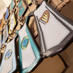 May include: A collection of white, teal, and gray keychains shaped like aprons with different designs. Some of the aprons have a heart shape with yellow and brown stripes, while others have a teal and yellow starburst design.