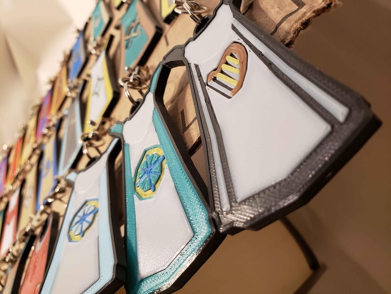 May include: A collection of white, teal, and gray keychains shaped like aprons with different designs. Some of the aprons have a heart shape with yellow and brown stripes, while others have a teal and yellow starburst design.