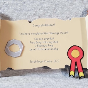 Op de afbeelding: Decoratieve scroll-vormige plaquette met de tekst "Gefeliciteerd! Je hebt de Huwelijksquest voltooid!" en andere tekst. Het heeft een zilveren ring en een rood lint met gele accenten. De plaquette is beige met bruine accenten.