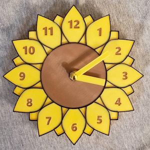 Op de afbeelding: Een gele en bruine wandklok in de vorm van een zonnebloem. De wijzerplaat is bruin, met gele bloemblaadjes aan de rand, elk gemarkeerd met een nummer. De wijzers zijn geel. Een decoratief item.