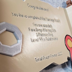 Op de afbeelding: Een 3D-geprinte rolvormige plaquette met de tekst "Gefeliciteerd! Je hebt de Huwelijksquest voltooid." Inclusief een zilverkleurige ring en een rood, geel en zwart lint. De plaquette is bruin en beige.