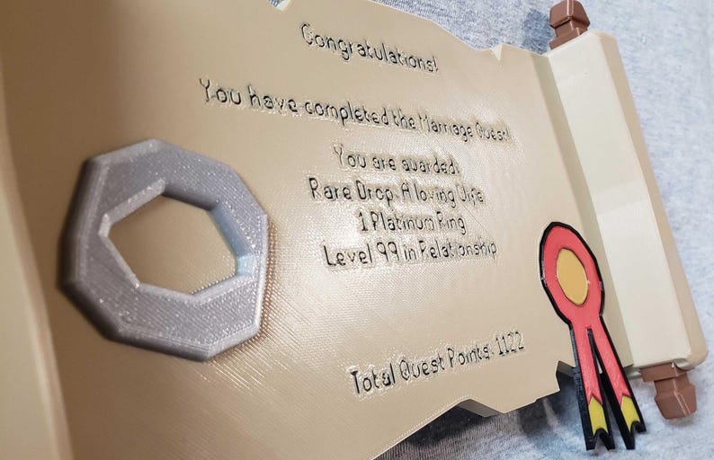 Op de afbeelding: Een 3D-geprinte rolvormige plaquette met de tekst "Gefeliciteerd! Je hebt de Huwelijksquest voltooid." Inclusief een zilverkleurige ring en een rood, geel en zwart lint. De plaquette is bruin en beige.