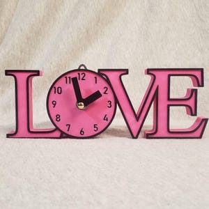 Op de afbeelding: Roze houten klok met het woord "LOVE" in roze letters. De wijzerplaat is roze met zwarte cijfers en wijzers. De letters hebben een zwarte omtrek.