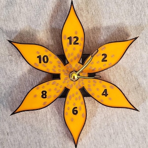 Pode incluir: Um relógio de parede em forma de flor com seis pétalas laranja e contornos pretos. O mostrador do relógio apresenta números pretos nas posições 2, 4, 6, 8, 10 e 12 horas. Os ponteiros do relógio são pretos e dourados.