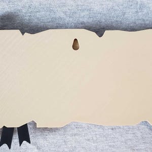 Op de afbeelding: Een beige, rolvormige plaquette met bruine decoratieve uiteinden en een zwart lint. De plaquette heeft een verweerde, verouderde uitstraling, met een klein gat om op te hangen. De rol is ontworpen om op perkament te lijken.