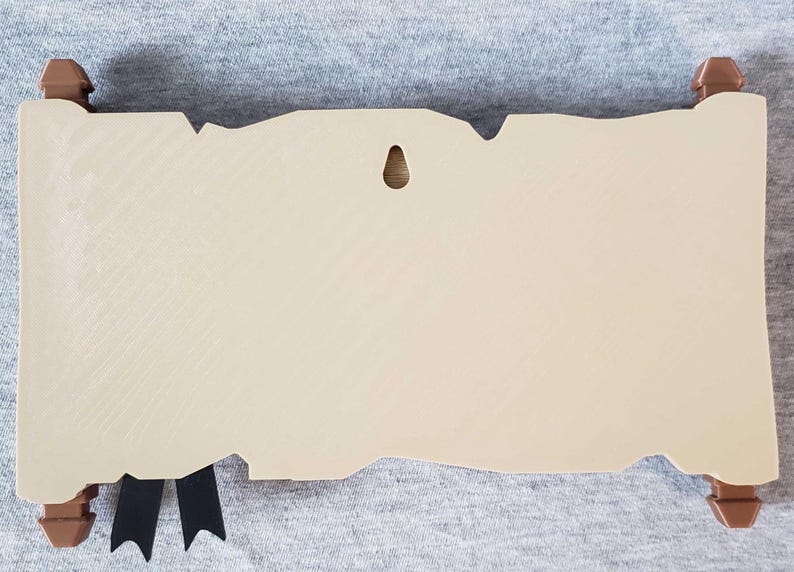 Op de afbeelding: Een beige, rolvormige plaquette met bruine decoratieve uiteinden en een zwart lint. De plaquette heeft een verweerde, verouderde uitstraling, met een klein gat om op te hangen. De rol is ontworpen om op perkament te lijken.