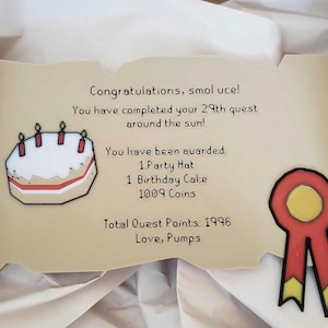Op de afbeelding: Een decoratieve rol met tekst en illustraties. De tekst luidt "Gefeliciteerd, smol uce! Je hebt je 29e zoektocht rond de zon voltooid!" en somt beloningen op. Bevat een verjaardagstaart en een rood lint.