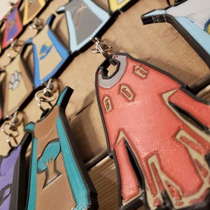 May include: A collection of colorful keychains featuring different designs. Each keychain is shaped like a cape with a unique pattern and color scheme. The keychains are made of a durable material and are perfect for adding a touch of personality to your keys.