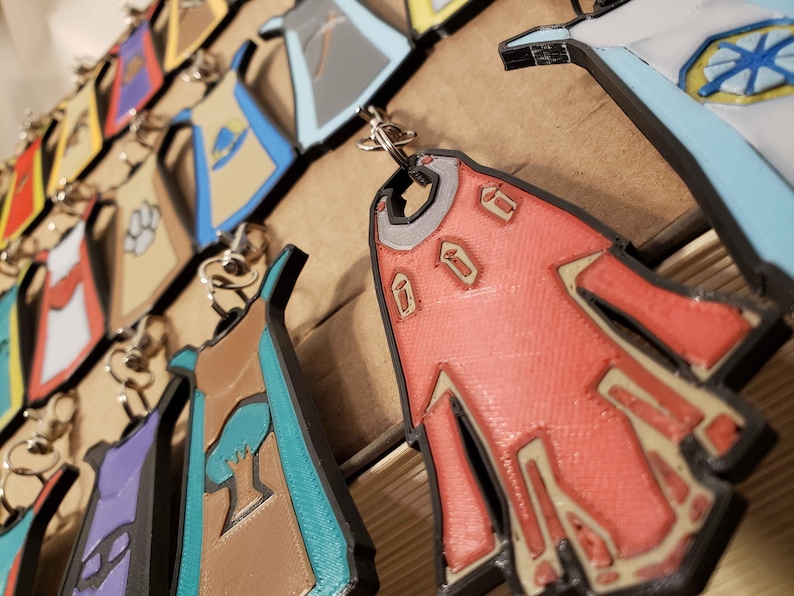 May include: A collection of colorful keychains featuring different designs. Each keychain is shaped like a cape with a unique pattern and color scheme. The keychains are made of a durable material and are perfect for adding a touch of personality to your keys.
