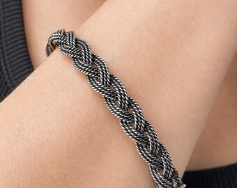 May include: Braided silver bracelet. The bracelet features a woven design with a secure clasp. The silver metal has a slightly oxidized appearance, giving it a vintage or antique look. The bracelet is shown on a person's arm.