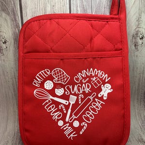 May include: Red quilted oven mitt with a white heart-shaped design featuring the words "butter", "flour", "milk", "sugar", "cocoa", and "cinnamon" along with illustrations of baking ingredients and tools.