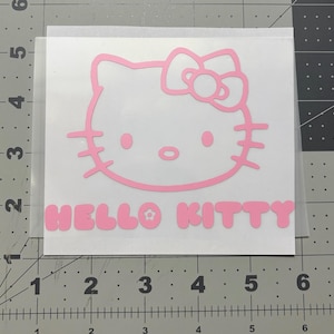 May include: A pink Hello Kitty decal. The decal features the iconic cartoon cat's face and the words "HELLO KITTY" in a matching pink font. The decal is on a clear backing.