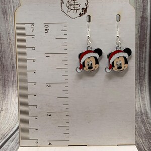 May include: Silver dangle earrings featuring a red and white Mickey Mouse wearing a Santa hat. The earrings are displayed on a wooden ruler with a measurement scale in inches and centimeters.