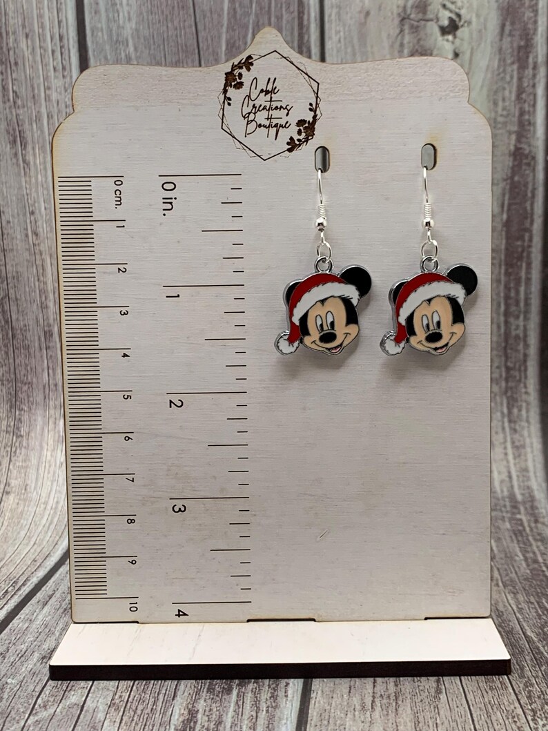 May include: Silver dangle earrings featuring a red and white Mickey Mouse wearing a Santa hat. The earrings are displayed on a wooden ruler with a measurement scale in inches and centimeters.