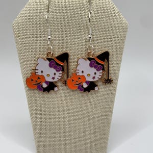 May include: A pair of silver dangle earrings featuring a white Hello Kitty character dressed as a witch holding a jack-o'-lantern. The earrings have a black witch hat, purple bow, and orange and black pumpkin.
