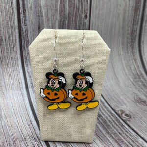 May include: A pair of dangle earrings featuring a cartoon character dressed as a pumpkin. The character is wearing a black hat and has a yellow face with a black nose and mouth. The pumpkin is orange with a green stem and a carved jack-o'-lantern face. The earrings are silver and have a hook closure.