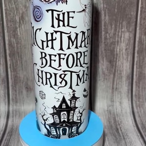 K&ouml;nnte beinhalten: Ein wei&szlig;er Becher mit einem Design aus "The Nightmare Before Christmas". Das Design zeigt Jack Skellington, Sally und ein Spukhaus. Der Text "The Nightmare Before Christmas" ist in einer stilisierten Schriftart dargestellt. Der Becher ist zylindrisch und steht auf einem blau-wei&szlig;en St&auml;nder.