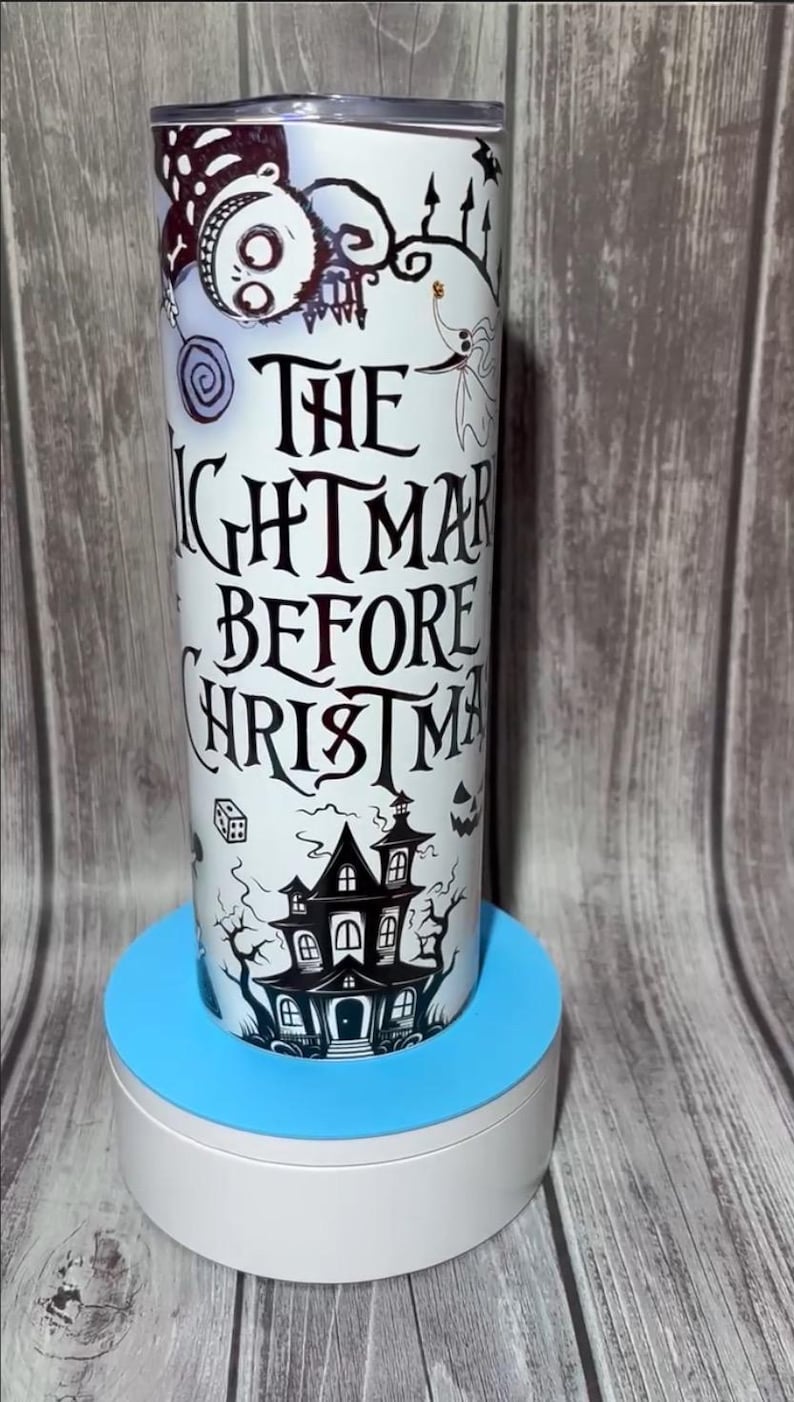 K&ouml;nnte beinhalten: Ein wei&szlig;er Becher mit einem Design aus "The Nightmare Before Christmas". Das Design zeigt Jack Skellington, Sally und ein Spukhaus. Der Text "The Nightmare Before Christmas" ist in einer stilisierten Schriftart dargestellt. Der Becher ist zylindrisch und steht auf einem blau-wei&szlig;en St&auml;nder.