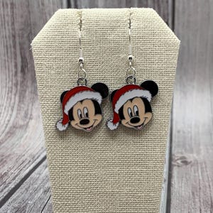 May include: A pair of silver dangle earrings featuring a cartoon Mickey Mouse wearing a red Santa hat. The earrings are on a beige fabric display stand.