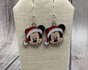 May include: A pair of silver dangle earrings featuring a cartoon Mickey Mouse wearing a red Santa hat. The earrings are on a beige fabric display stand.