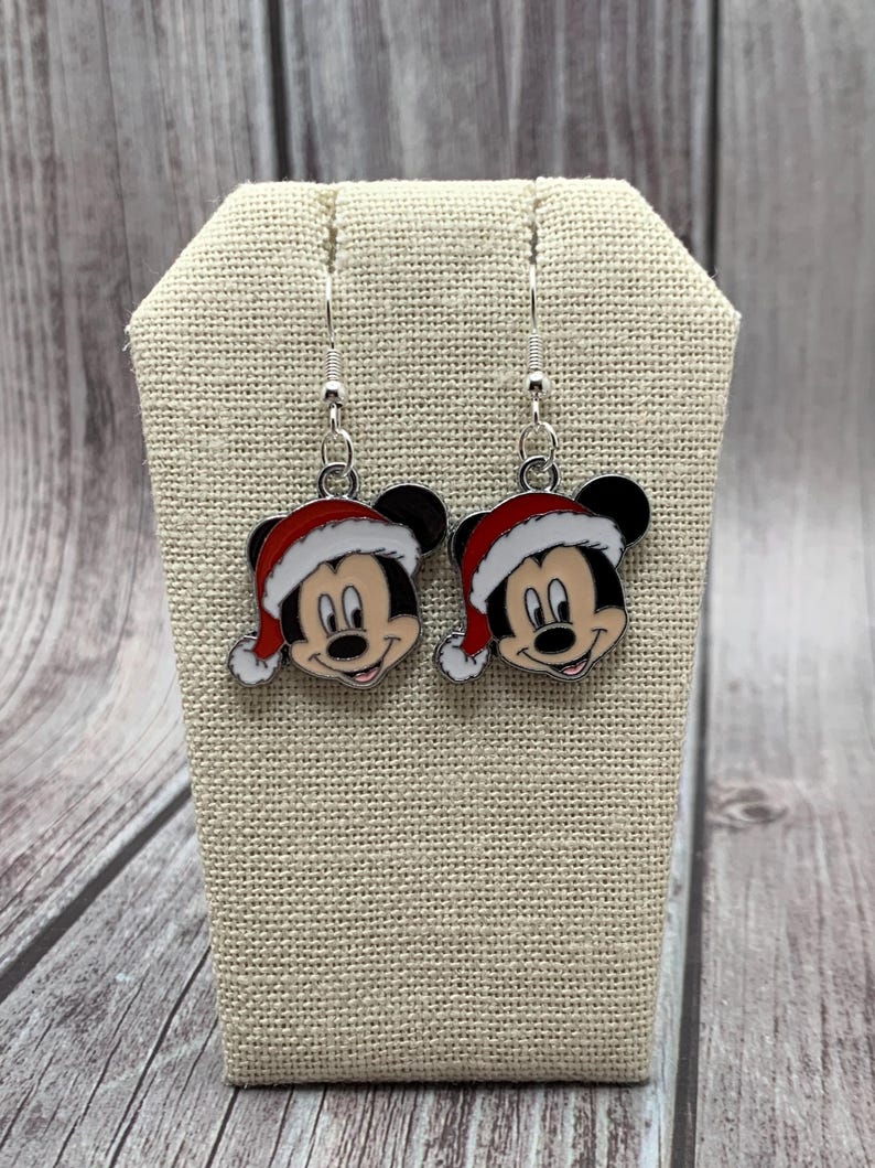 May include: A pair of silver dangle earrings featuring a cartoon Mickey Mouse wearing a red Santa hat. The earrings are on a beige fabric display stand.