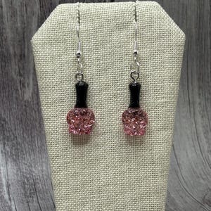 May include: A pair of dangle earrings featuring pink glitter nail polish bottle charms with black tops and silver hooks.