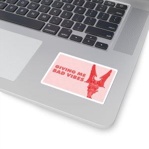 Könnte beinhalten: Ein rosa Aufkleber mit einer roten Illustration einer Gargoyle mit Flügeln und dem Text "Giving Me Bad Vibes".