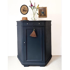 Könnte beinhalten: Ein dunkelblauer, antiker Schrank mit einer Tür und einer Schublade. Ein kleiner Strohbesen hängt an der Tür. Ein Strohhut, eine Vase mit Blumen und zwei gerahmte Bilder stehen auf dem Schrank.