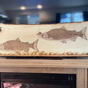 May include: A wood burned art piece featuring two salmon on a natural wood slab. The salmon are detailed with wood burning and are set against a background of small wood burned rocks.